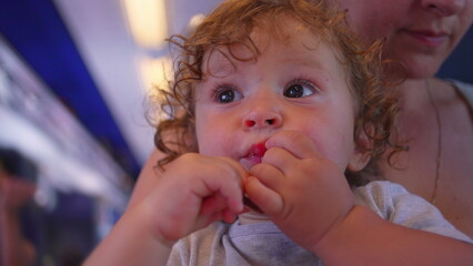 Curly-haired toddler bites into chocolate bar while looking up, seated on mother&rsquo;s lap inside moving train with cabin lights glowing