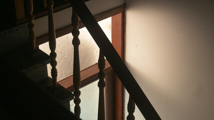 Light comes through a window above a door into a dark room with stairs. Door with light behind it near the railing of the stairs