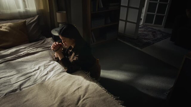 View from above of religious young woman praying with rosary beads while sitting on floor with elbows on edge of double bed in dark room