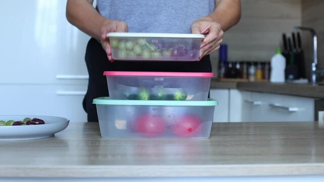 A person stacks three plastic containers filled with fresh produce on a kitchen counter, showcasing food storage and organization.