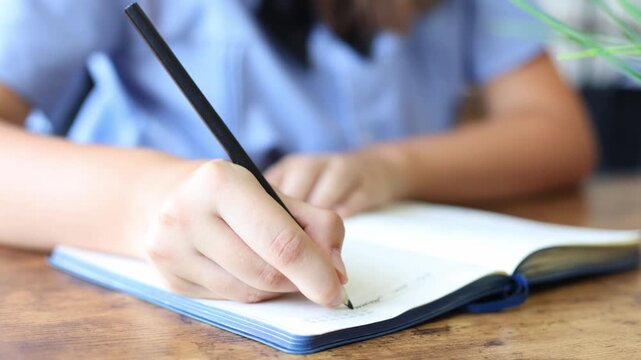 A student in a light blue shirt writes diligently in a notebook with a black pencil, focused on her schoolwork at a wooden desk.