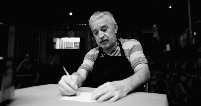 Older caf&eacute; owner struggles with calculations at table conveying uncertainty financial confusion mounting pressure and fear of collapse for small neighborhood business, monochrome