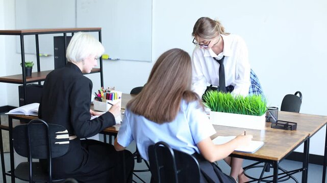 Three women collaborate at a desk in a modern office, discussing work with notebooks and pens, fostering a focused team environment.