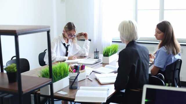 Three women collaborate at a sunlit desk, discussing notes and reviewing work in a modern, productive office or classroom environment.
