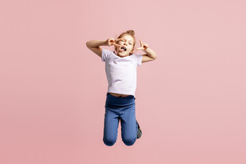 Playful girl jumping in air making silly faces with tongue out on pink background