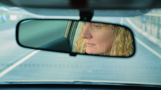 Driver's perspective of a woman's face reflected in the rearview mirror. Motion blur and cool tones create a pensive mood, perfect for concepts of travel, journey, road trips, and introspection