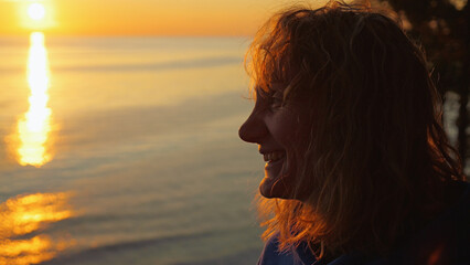 Fototapeta premium Profile of a smiling woman with messy hair, backlit by a warm, golden sunset. She gazes at the sun's reflection on the water, feeling content and peaceful. Serene travel and lifestyle concept