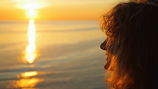 Profile silhouette of a woman watching a golden sunset over the ocean. Warm, serene lighting highlights her hair. The mood is peaceful, contemplative, hopeful. Soft focus background with copy space
