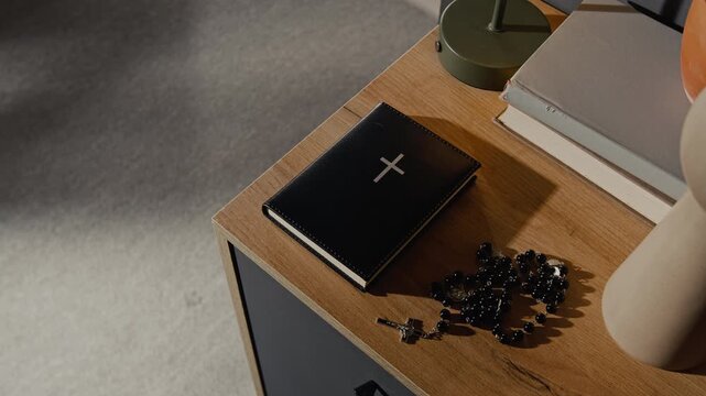 Close-up view of holy bible in leather cover and black rosary beads with cross next to books and decor items on wooden top of cabinet