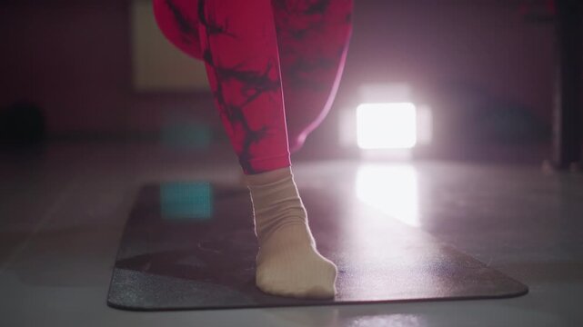 asian trainer balancing on mat in lunge, spotlight backlight silhouette, singleleg strength work in pink leggings and socks, controlled knee alignment, core engagement and steady breath, gritty