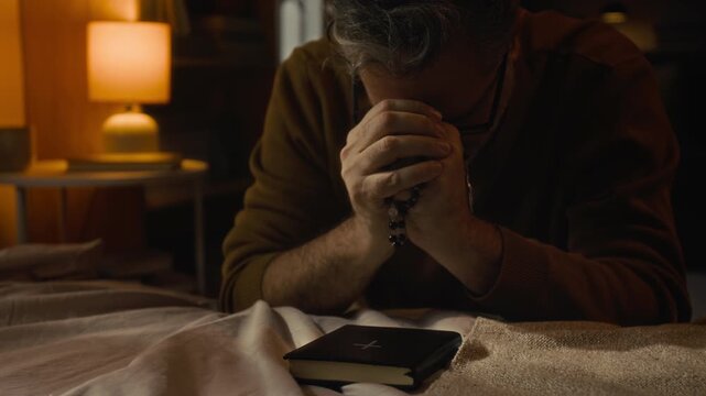 Grey-haired man leaning forward with his hands clasped together in prayer while holding rosary, with closed bible placed on bed in front of him in dimly lit room