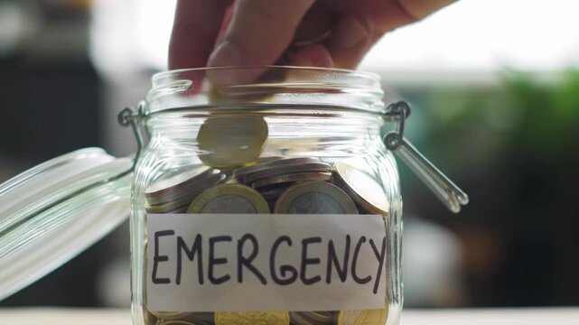 A pristine clear glass jar with 'EMERGENCY' label and accumulating gold/silver coins. A well-groomed hand drops a gleaming gold coin. Hopeful financial security concept