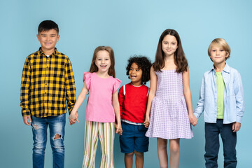 Fototapeta premium Group of smiling diverse children holding hands looking at camera posing isolated on blue background