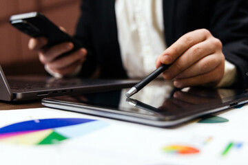 Business professional using smartphone and stylus on tablet while analyzing financial charts and graphs on printed documents in modern office setting