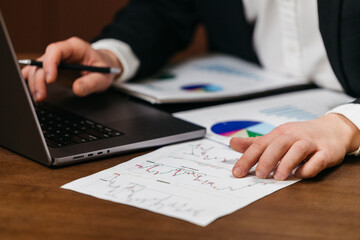 Business professional analyzing financial reports with a laptop and printed charts on a wooden desk, pen in hand, focused on data and decision-making. Concept business analysis, finance, office work