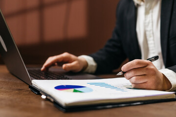 Business professional analyzing financial data on a laptop while taking notes with a pen on a report featuring colorful charts and graphs on a wooden desk
