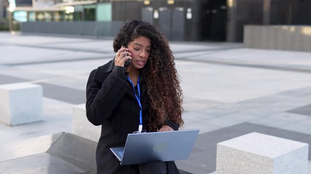 Young, accomplished businesswoman with elegant curls multitasks with her laptop and smartphone as she sits in a bustling urban square