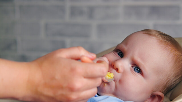 Tight close-up of a blue-eyed baby being fed yellow puree from a spoon by a parent. The shallow depth of field focuses on the infant's face. Authentic moment of childcare, nutrition, and development