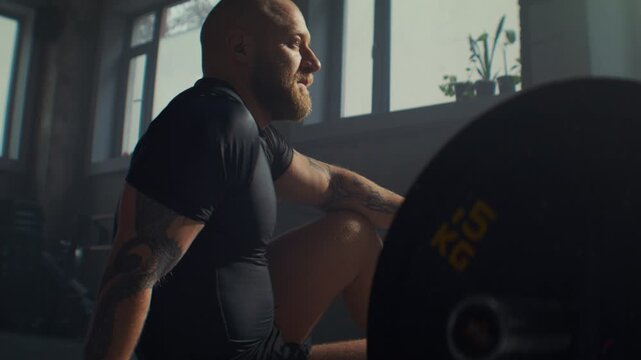 Tired sportsman rests sitting near barbell on gym floor side view