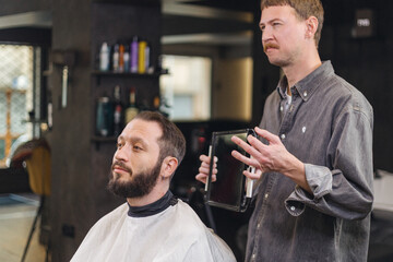 Barber showing haircut result to male client using mirror in barbershop. Professional consultation,...