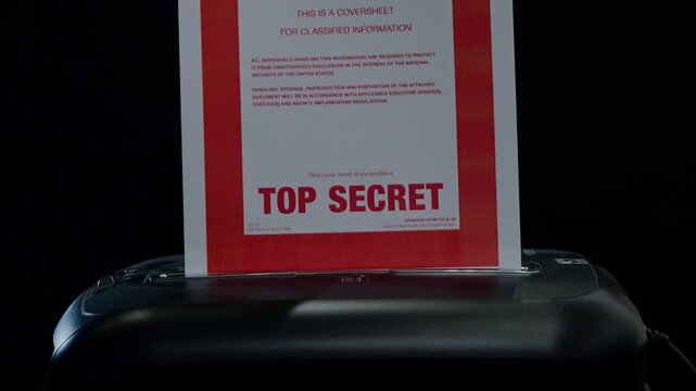 A coversheet to a confidential document is fed into a paper shredder. Subdued lighting. Suggestive of spying, espionage, or cover-up.