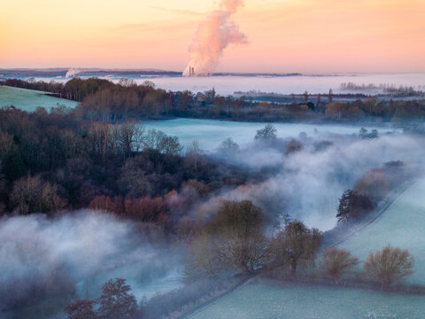 Aerial view of mist-laden fields and frosty tree lines meeting the distant Didcot Power Station's plumes under a pastel sky, Wittenham Clumps, Oxfordshire, England.