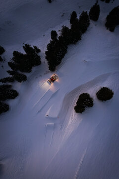 Aerial view of snow groomer navigates the snowy slopes near dark, casting a warm glow against the cold, white landscape, Val-d'Illiez, Valais, Switzerland.