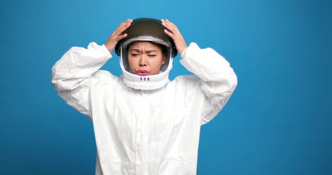 Sad hispanic woman wearing helmet and white spacesuit having headaches, isolated on blue background