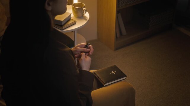 Midsection shot of brunette woman holding rosary beads and taking holy bible during morning prayer in home setting next to white bedside table with books, mug and lamp