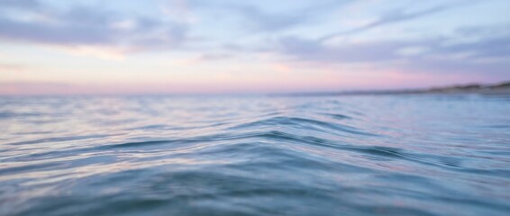 Calm blue sea water surface with gentle ripples in a close-up, low-angle view against a beautiful blurred pastel sunset sky with soft clouds