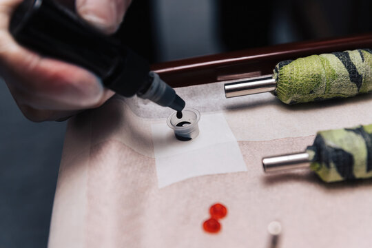 Tattoo artist pouring black ink into a small cup on a tray, with tattoo machines nearby, showing preparation process.