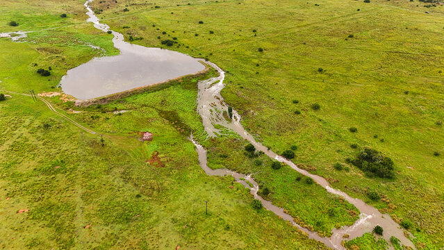 Aerial view of a small dam overflowing into a winding stream cutting through a vibrant green landscape, Vaalwater, Limpopo, South Africa.