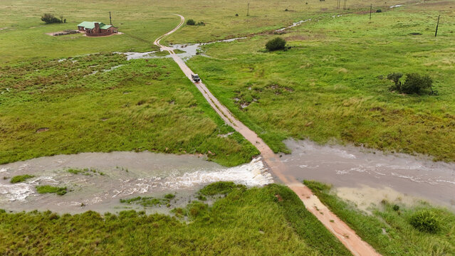 Aerial view of a car splashes through a flooded dirt road cutting through vibrant green fields, contrasting against the distant house, Vaalwater, Limpopo, South Africa.