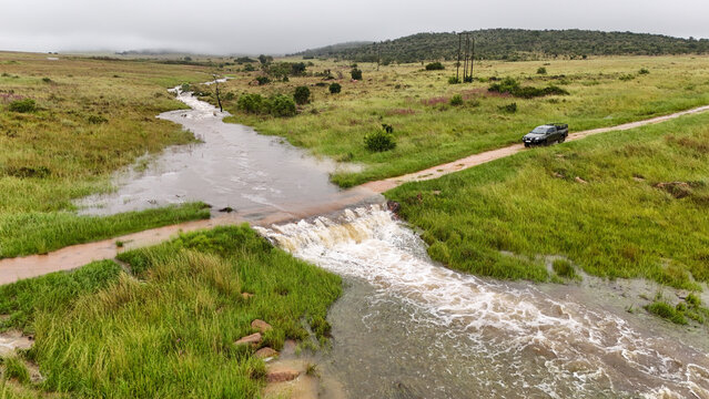 Aerial view of a vehicle cautiously crossing a submerged road amidst verdant grasslands and turbulent waters, Vaalwater, Limpopo, South Africa.