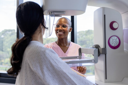African American tech in pink scrubs guiding Asian patient in gown at mammogram unit with shield