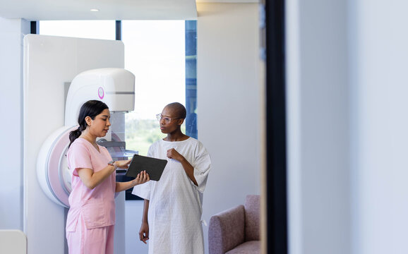 Diverse female tech in pink scrubs consulting patient in gown at mammography machine, using tablet