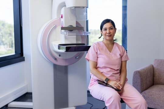 Female rad tech in pink scrubs sitting beside mammography unit with paddle and tablet, copy space