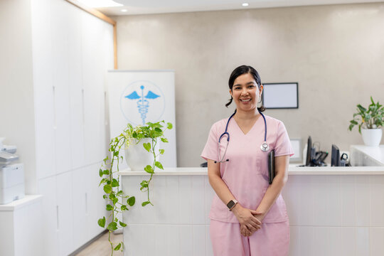 Female nurse in pink scrubs standing behind white reception desk holding clipboard and stethoscope