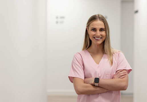 Adult female clinician standing arms crossed smiling in clinic corridor in pink scrubs smartwatch