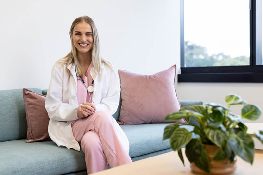 Female doctor sitting on light teal sofa in clinic, wearing pink scrubs and stethoscope, smiling