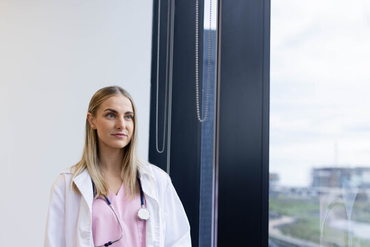 Female clinician looking out clinic window, blinds in view, white lab coat pink scrubs, stethoscope