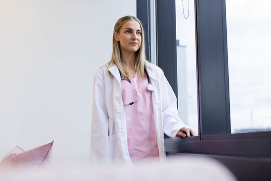 Adult female nurse standing in clinic near tall window, gazing, wearing pink scrubs and stethoscope