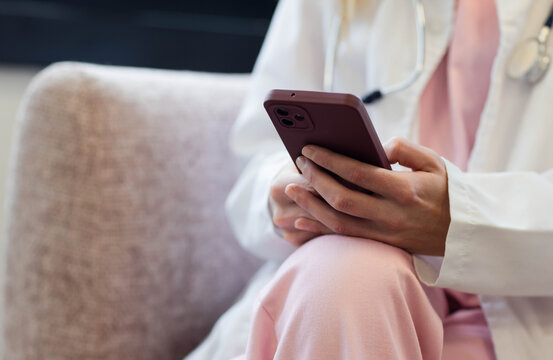 Hands are holding maroon smartphone and typing, wearing white coat and stethoscope on beige sofa