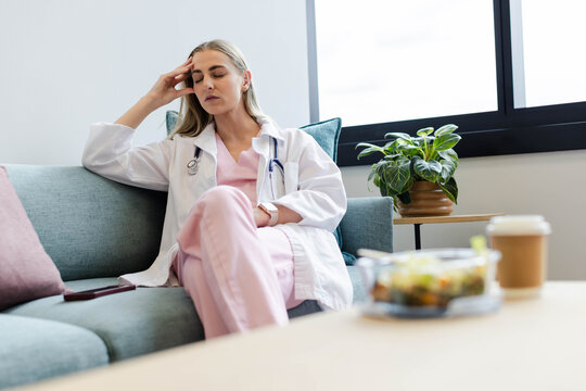 Female in scrubs sitting on teal sofa in lounge rubbing temple, stethoscope smartphone, copy space