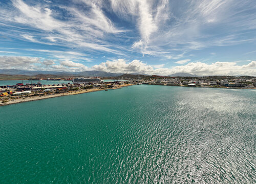 Aerial view of the shimmering turquoise harbor contrasting with the industrial docks and the distant mountain range under a bright, cloud-strewn sky, Noumea, South Province, New Caledonia.