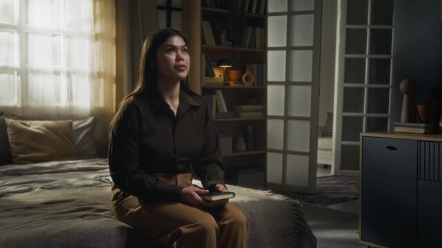 Thoughtful young woman wearing dark brown shirt and sand-coloured pants talking to God sitting on edge of bed with bible book