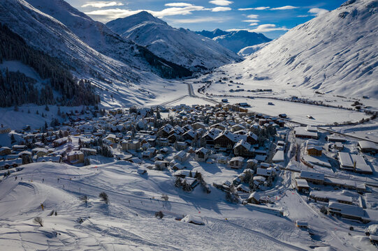 Aerial view of a snow-covered village nestled in a valley, surrounded by towering mountains under a clear blue sky, Andermatt