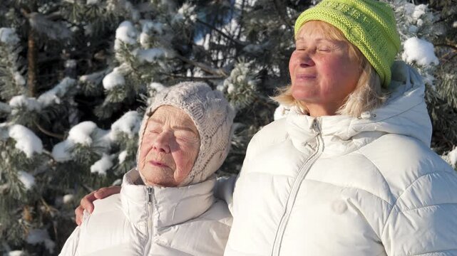 Two elderly women relatives in white jackets sharing joyful moment outside, taking sunbath. Old mother and adult daughter clad in white garments enjoying serene winter day amidst snowy pine trees