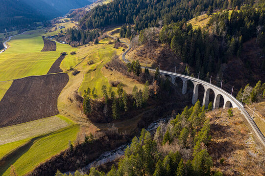Aerial view of a stone railway bridge curving through green and brown fields, contrasting with dark forest patches, Bergun Filisur, Grisons, Switzerland.