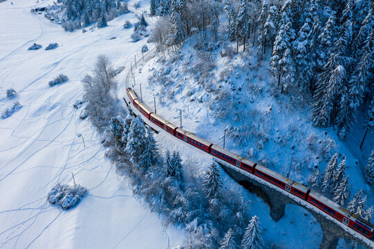 Aerial view of a red train slicing through the snowy landscape, contrasting against the white expanse and dark evergreens, Bergun Filisur, Grisons, Switzerland.